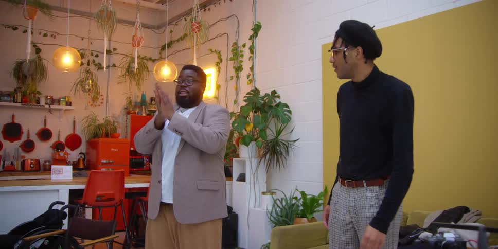 Two black men stand in a room talking and acting out