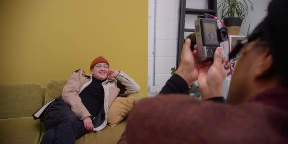 A man photographing a man sitting on the yellow sofa