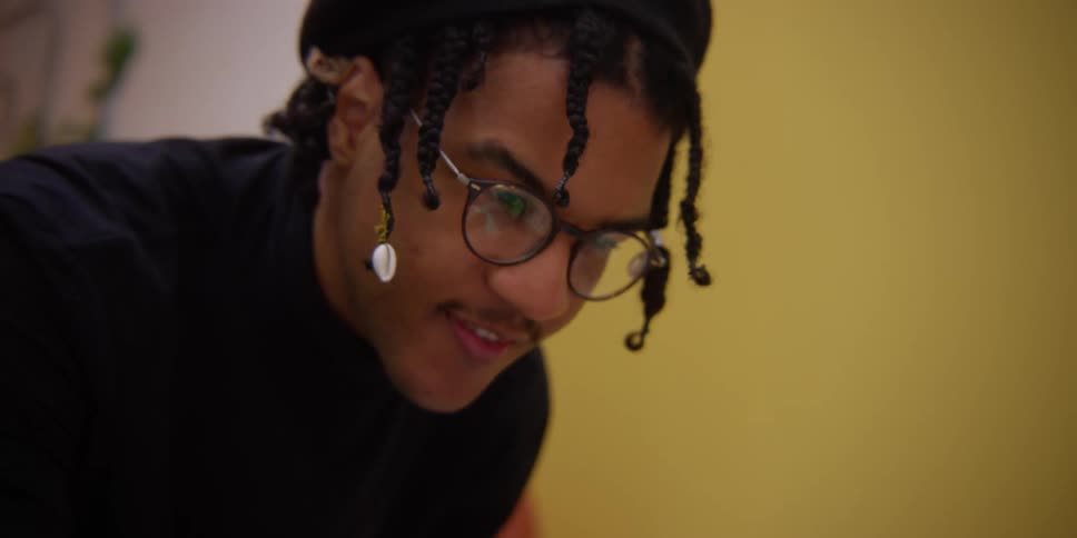 A person in a black hat and curly dreads in front of a yellow background