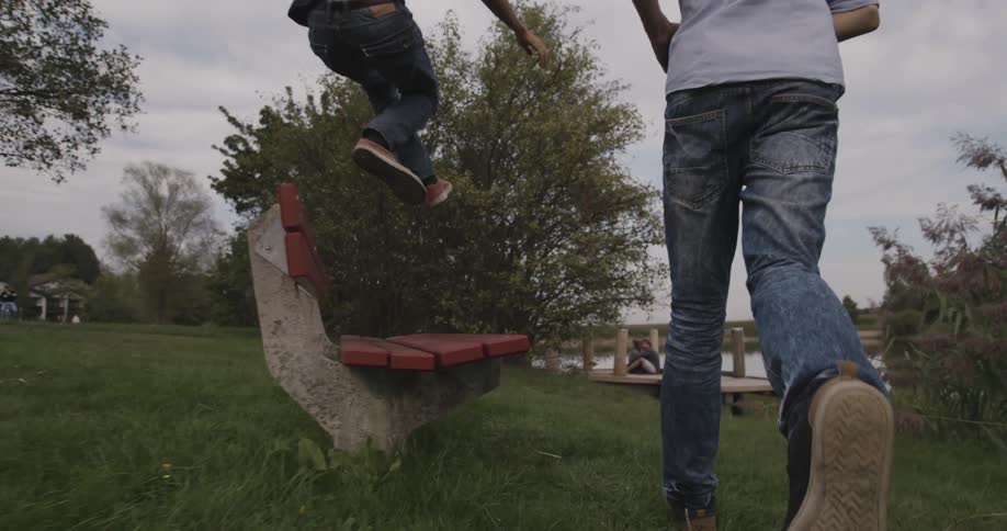 Two kids run over a grass field one jumps over a bench