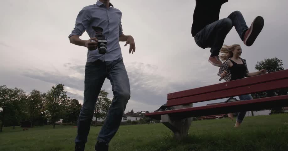 Young people run through a field and a young man jumps over a wood bench