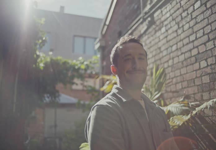 A happy man smiles standing outside near a brick wall with green plants