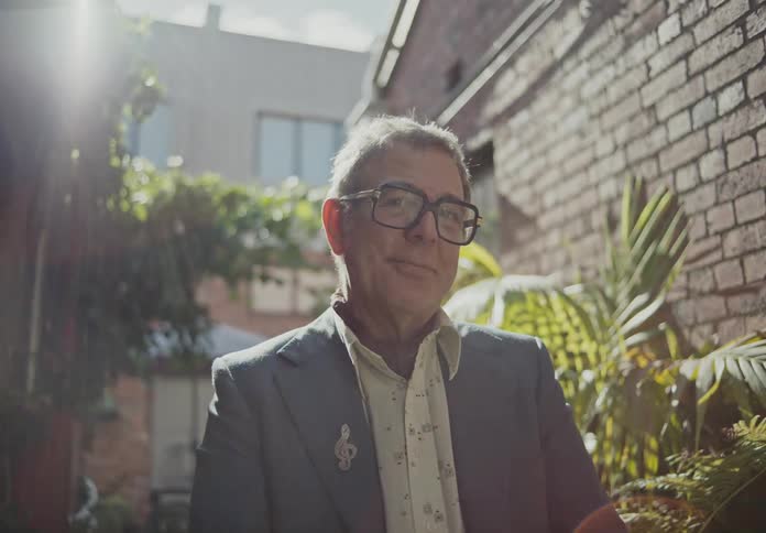A man wearing glasses stands near a wall with plants and buildings in the background