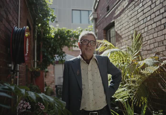 An older white man stands in a cluttered alley while smiling