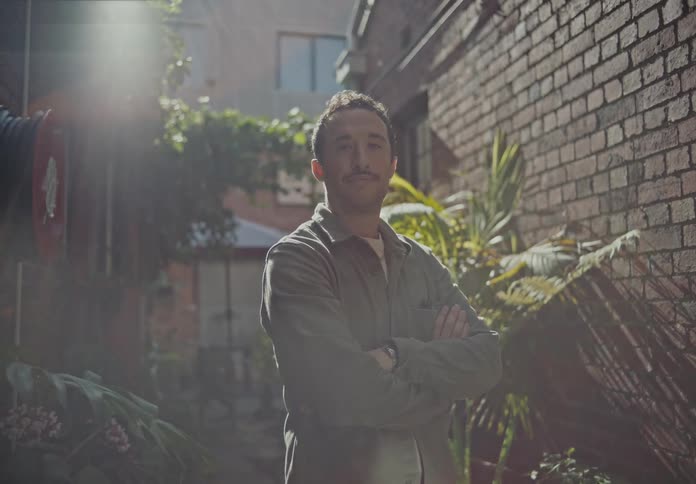 A person is standing with arms crossed in a narrow alleyway surrounded by brick walls and green plants