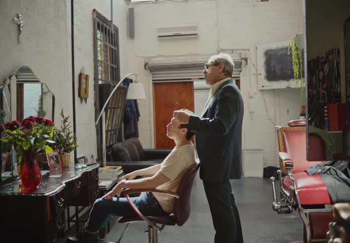 A barber is standing talking to a man who is sitting and looking in the mirror