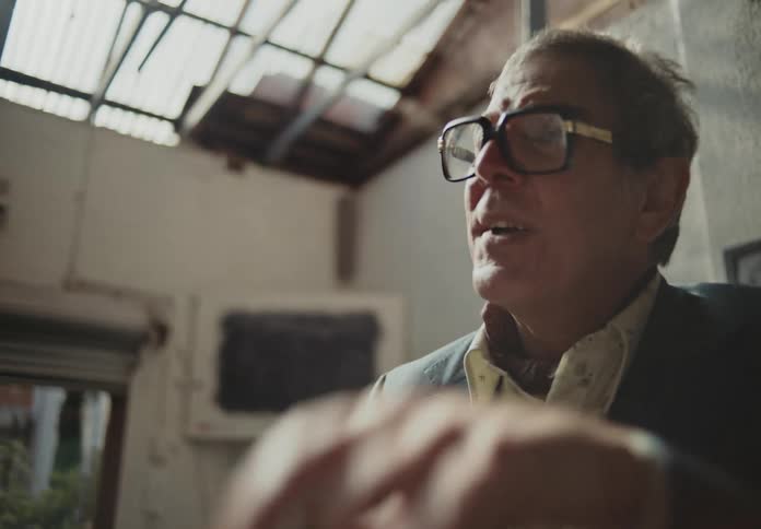 An old man in reading glasses and formal clothes is talking to someone in front of him and interacting with an object