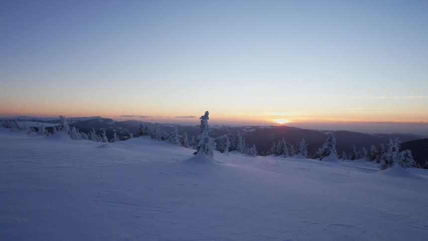 Snow covered plain overlooks frozen conifer forested valley as sun sits on horizon over mountains