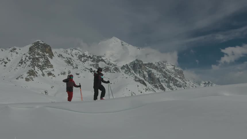 Two people with ski equipment are walking on a snowcovered surface and there is a mountain behind them
