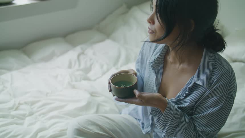 A young women drinks out of a cup while sitting on a bed