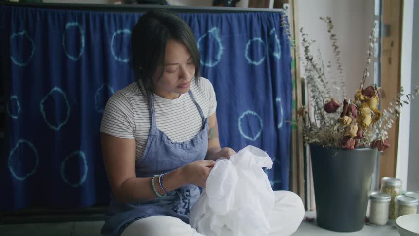 A young woman sits in a room stitching a piece of fabric