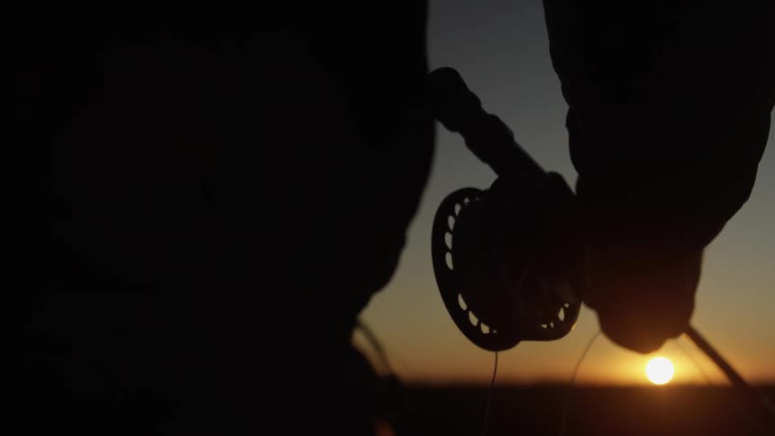 Slow motion frame of the silhouette of a man adjusting the fishing rod line with a sunset on the horizon