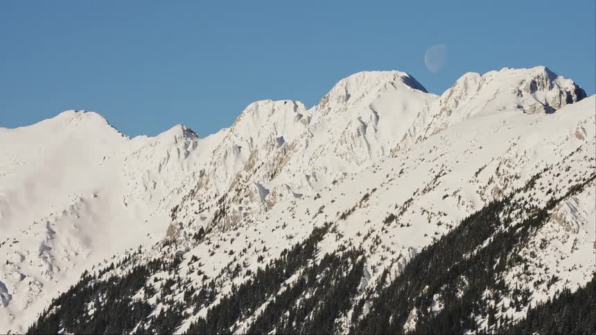 Mountain range covered by snow on a day with a cloudless blue sky and a view of the moon