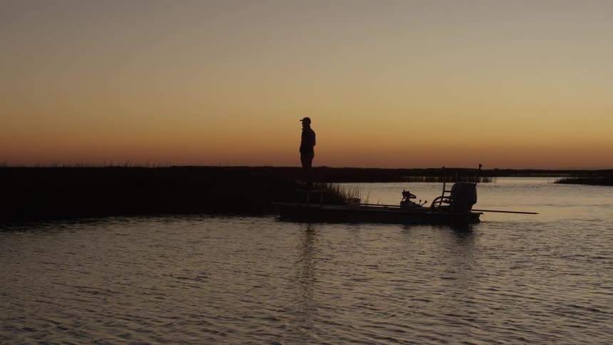 The silhouette of a person walking out of a boat onto dry land and stopping to admire the orange dawn environment