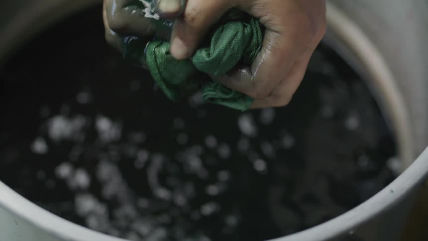 A pair of hands squeezes a dark green dye from cloth above a white bucket
