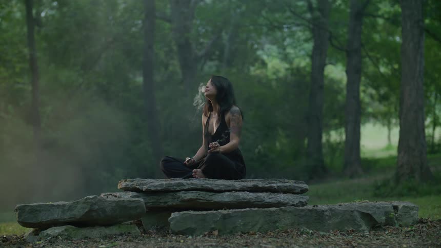 A woman sits on rocks in a forest and smokes
