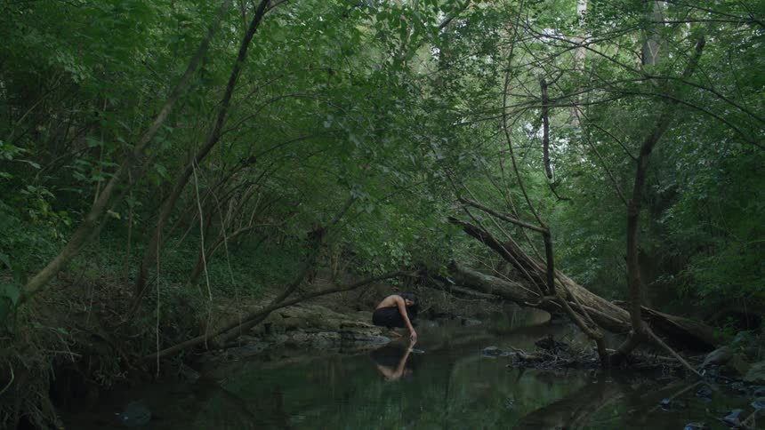 Woman crouching in front of a river with her reflection reflecting in the middle of a dense forest