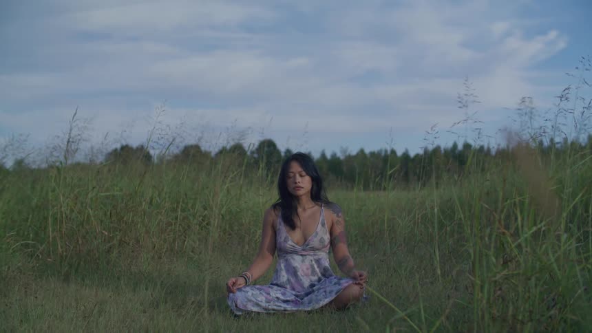 Woman sitting in a yoga pose with closed eyes amidst a field with tall grass