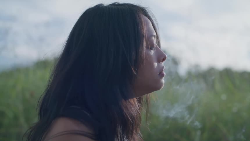 A woman smoking in a grassy field with an overcast sky