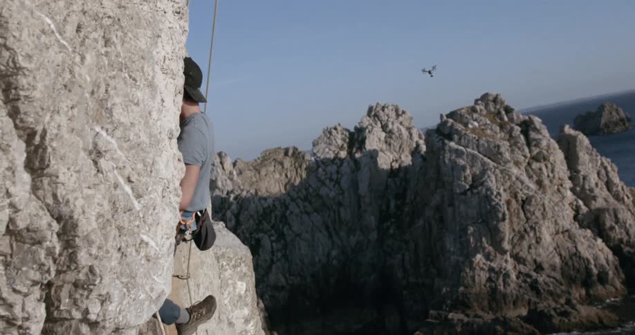 A man with a backwards cap is descending a rock using a rope tied around his waist in an area with many rocks