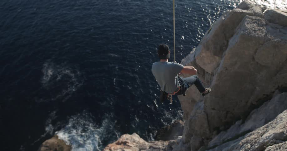 Male rock climber dangles over ocean on rope attached to seaside cliff