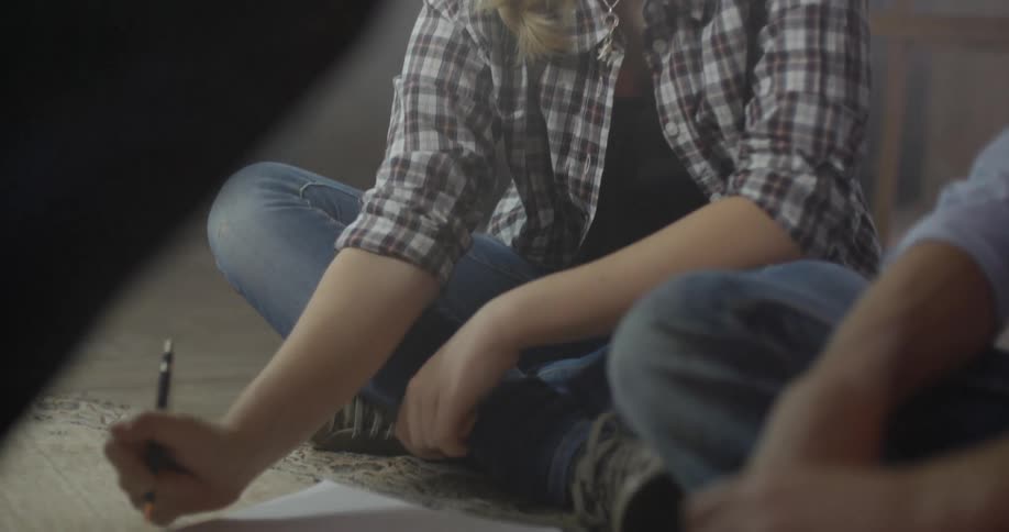 A woman sitting on the floor with others is holding a pen near a writing pad