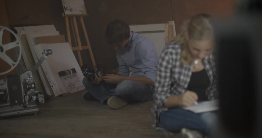 Two people are sitting in a studio one is drawing one is using a camera