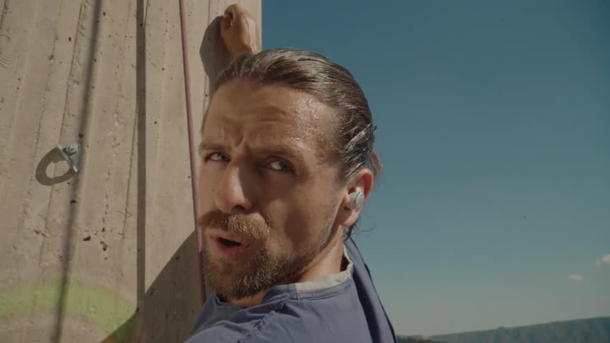 A white man with an earbud leans against a concrete wall