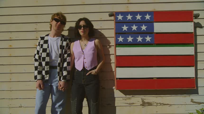 Two models side by side posing with sunglasses and a wooden background with the united states flag next to them