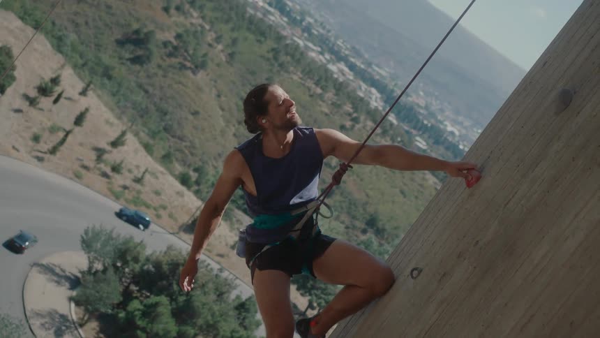 A man is rock climbing while a rope is attached to him for safey as cars are seen going by on the floor