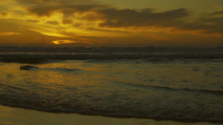 An ocean spills small waves onto a beach at dusk with the sunset behind it