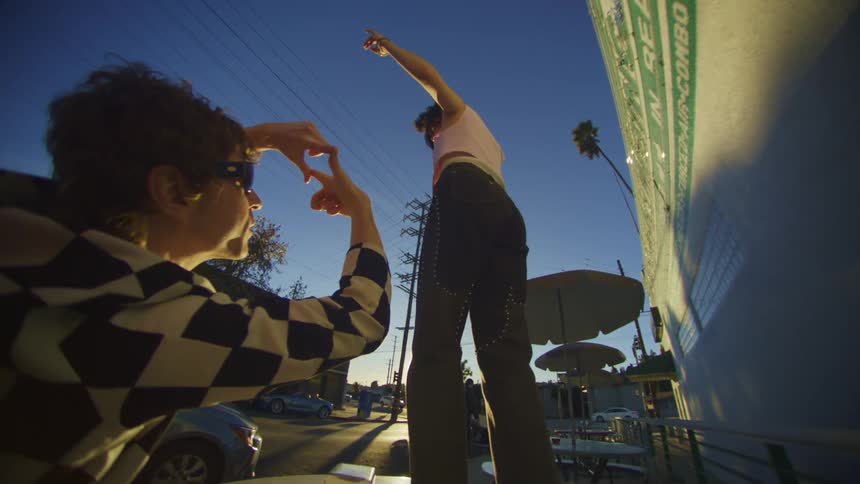 Man mimics camera with his hands while woman twirls near outdoor cafe as sun sits near horizon