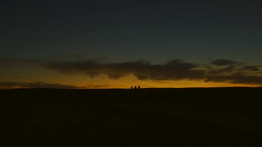Landscape of a grassland with the silhouette of 3 people in a dark reddish dawn sky