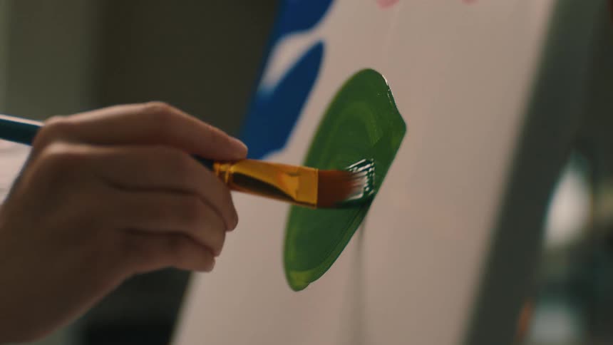 Closeup of a womans hand passing a brush with green paint on a painting