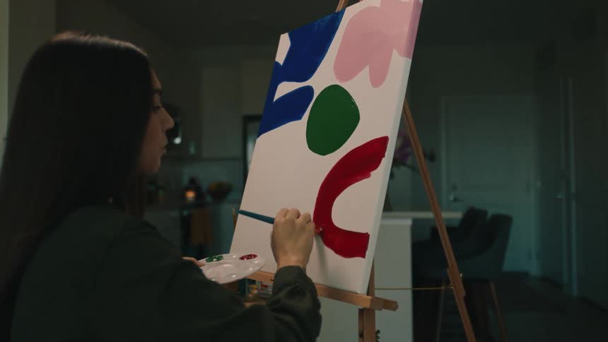 Female artist paints red shape onto canvas on easel set up in room near kitchen