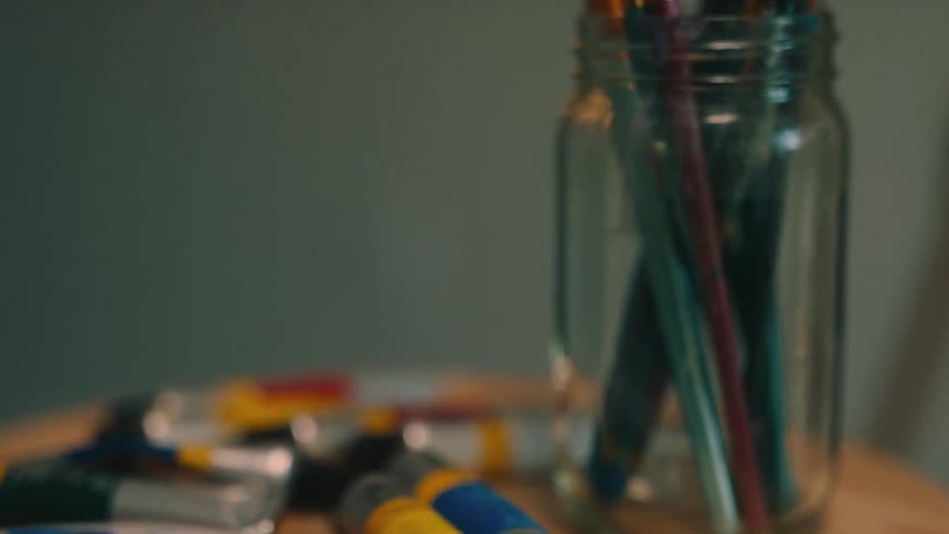 Art supplies including paintbrushes and paints sit on wooden stool near easel