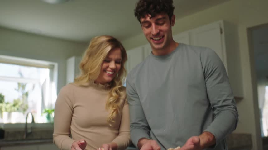A man and a woman are standing preparing cookies and the woman kisses the man on the cheek and they both smile
