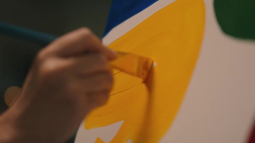 Hand of a female painter indoors holding a brush and spreading yellow paint over a white canvas
