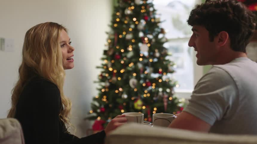 Couple sitting on the sofa holding cups with a decorated christmas tree in front
