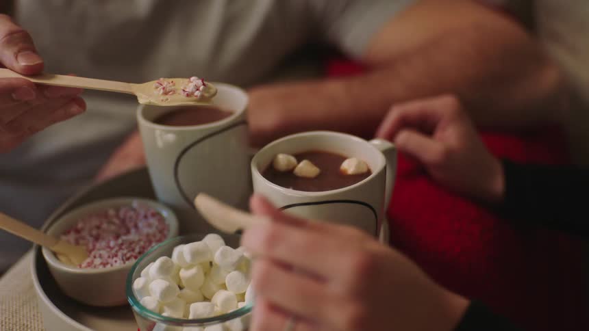 Two people on couches prepare hot chocolate with marshmallows and candy