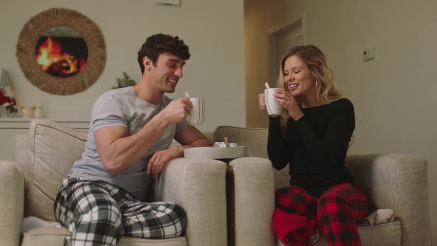A man and a woman sitting on a chair in the living room drinking a cup of tea as they talk and smile at each other