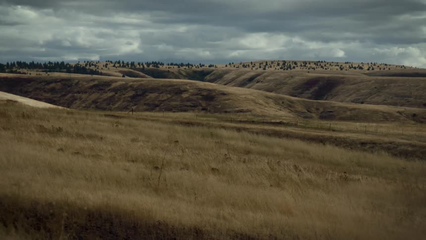 Semi truck passes between grass covered hills under dark clouds