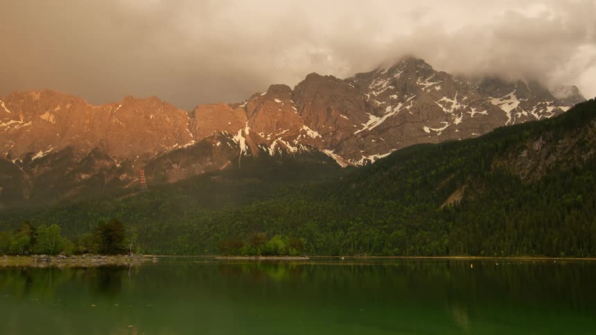 A serene lake reflects the mountains and forest under a cloudy sky