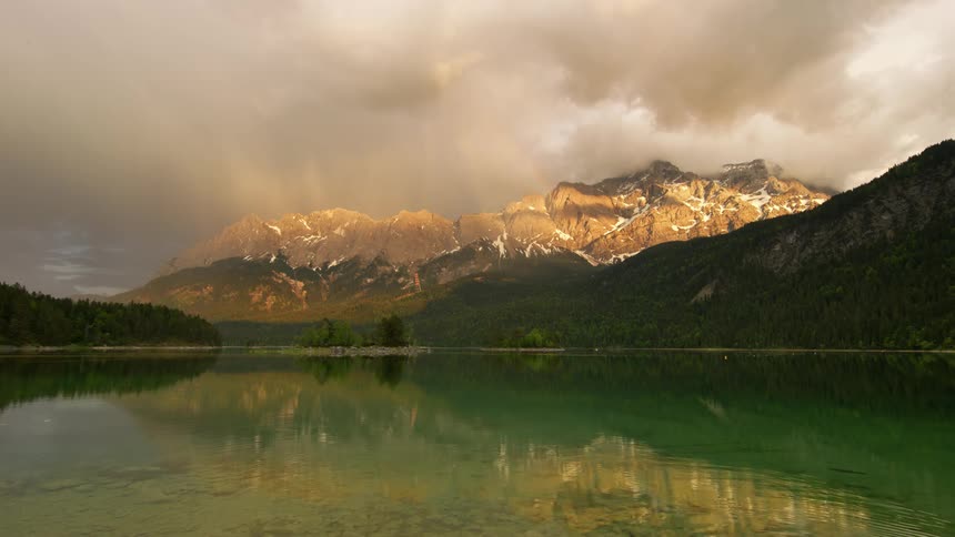 Landscape of a greenish lake surrounded by mountain ranges with mossy trees and snow on a cloudy day