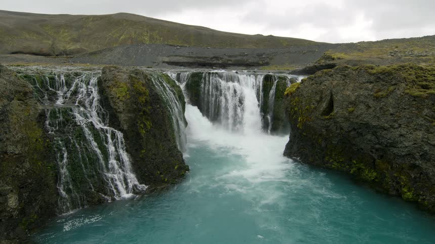 Water flows down multiple waterfalls near mountain into blue colored river on an overcast day