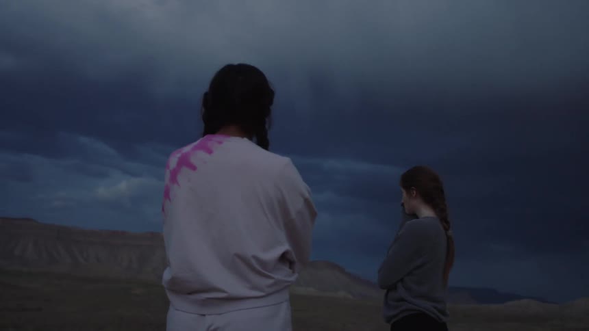 Two women standing with their backs turned in a desert region and one of them turns around