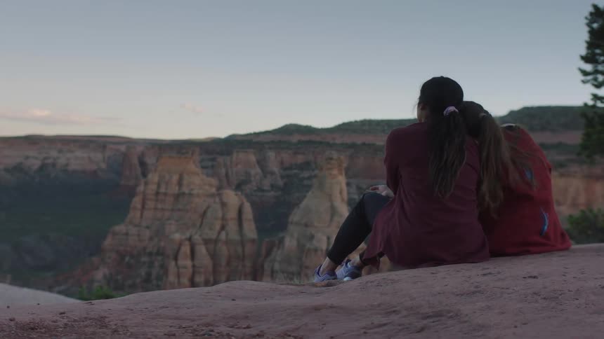 Two woman outdoors in nature sitting next to each other at the peak of a mountain leaning their heads against each other during day time