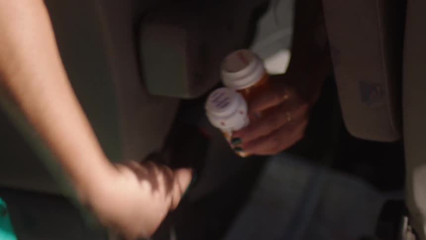 Woman in the front drivers seat picking up some medicines that are on the floor in the back seat