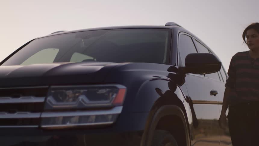 Woman gets out of car and walks past the hood to stand silhouetted against the bright sun