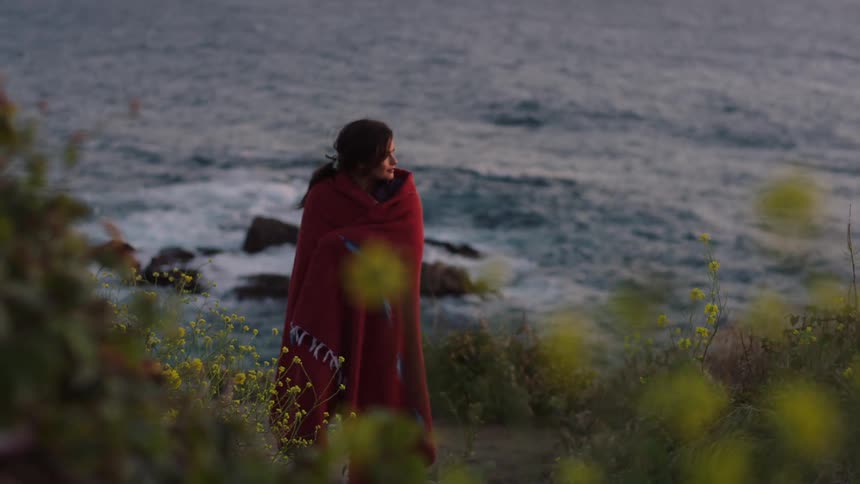 A young woman in a red cloak walks near a coastal area